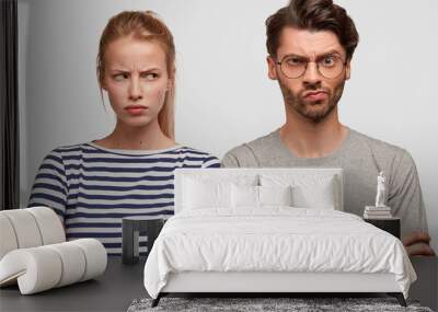 Indoor shot of grumpy European couple have displeased expressions after quarrel, don`t speak to each other, look angrily, keep hands crossed can`t find common solution, isolated on white wall Wall mural